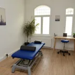 Ein heller Therapieraum mit einer Behandlungsliege, großen Fenstern und Physiotherapie Equipment.
